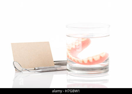 Close up di dentiere in un bicchiere di acqua e strumento dentale con avviso sulla carta backgrounda bianco Foto Stock