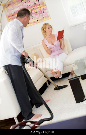 L uomo aspira, donna seduta su un divano a leggere un libro Foto Stock