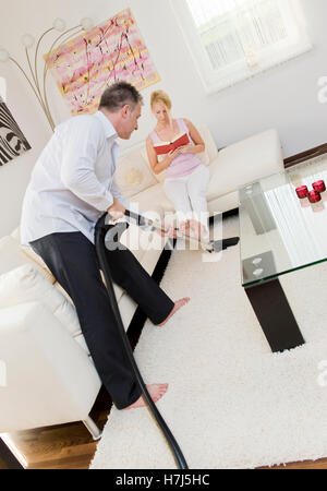 L uomo aspira, donna seduta su un divano a leggere un libro Foto Stock