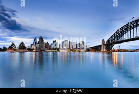 La città di Sydney CBD landmarks attraverso Harbou da case a ponte ad arco ora blu del tramonto quando le luci riflettono ancora in acque Foto Stock