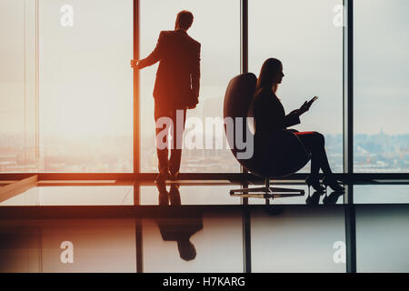 Silhouette di imprenditore in piedi accanto alla finestra grande, guardando sfocato cityscape esterno, il suo collega di sesso femminile con la compressa Foto Stock