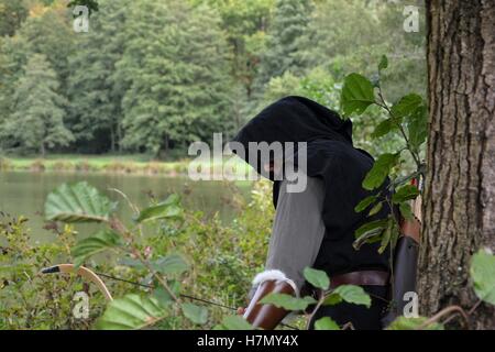 Arciere medievale con cappa nera sta nascosto dietro albero nel lago con curva tesa Foto Stock