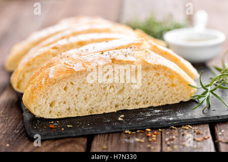 Freschi Fatti in casa ciabatta pane affettato su pietra ardesia bordo, olio di oliva e rosmarino su sfondo di legno Foto Stock