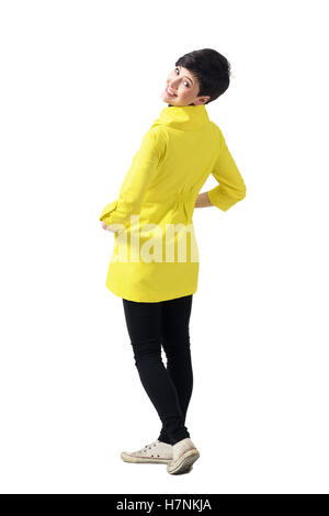 Vista posteriore del giovane giocosa donna che indossa cappotto giallo girando e sorridente alla fotocamera. Corpo pieno lunghezza ritratto isolato su bianco Foto Stock