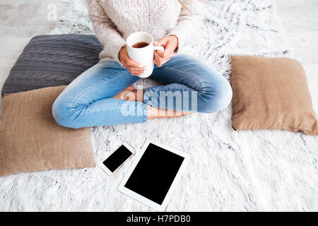 Immagine ritagliata di una donna azienda tazza di tè mentre è seduto sul tappeto con schermo vuoto di tablet e smartphone in ambienti interni Foto Stock