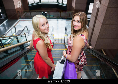 Due belle giovani donne che indossano abiti da shopping insieme e cavalcare un Escalator in un centro commerciale complessa e guardando indietro alla fotocamera Foto Stock