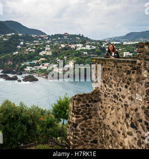 Una donna sta ponendo da un antico muro di pietra del castello aragonese affacciato sulla costa dell'isola di Ischia; Campania, Italia Foto Stock