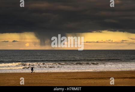 Silhouette di una persona e cane a piedi sulla spiaggia al bordo dell'acqua sotto nuvole di tempesta con strisce di pioggia e Un cielo al tramonto in lontananza oltre ... Foto Stock