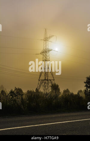 Alta tensione La torre di trasmissione vicino strada rurale. Energia elettrica sul filo di nebbia di mattina sfondo. Foto Stock