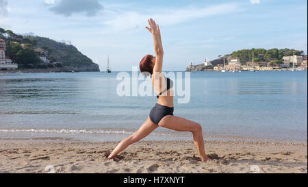 Una donna a praticare yoga su una spiaggia in Maiorca. guerriero 1 pongono mostrato. Foto Stock