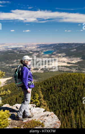 Escursionista femmina sulla scogliera rocciosa bordo affacciato sulla valle sottostante con dolci colline, lago, cielo blu e nuvole, Kananaskis Paese; Alberta, Canada Foto Stock