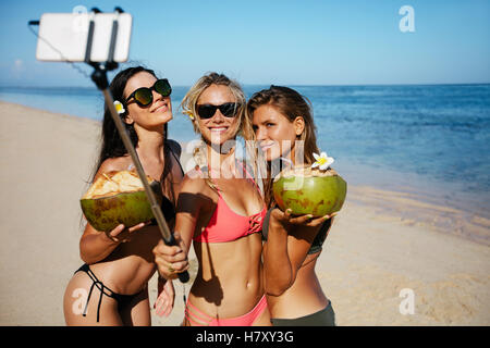 Gruppo di giovane donna azienda noci di cocco e tenendo selfie sulla spiaggia. Tre giovani donne in costume da bagno sulla spiaggia prendendo self por Foto Stock