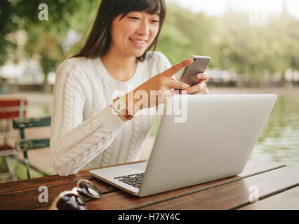 Colpo di felice donna asiatica utilizza lo smartphone a outdoor coffee shop. Donna seduta al tavolo del bar con il computer portatile all'aperto utilizzando mobil Foto Stock