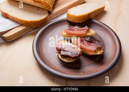 Vista ravvicinata di saporite patate al forno con fette di pancetta in argilla marrone piastra su sfondo di legno. Fette di pancetta affumicata Foto Stock