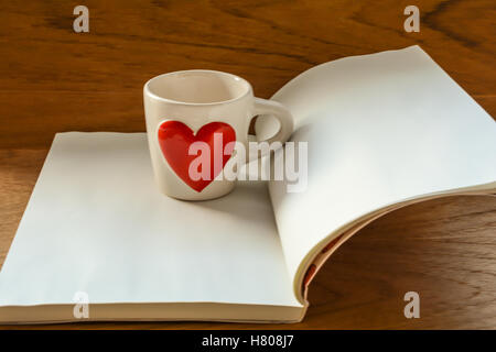 Tazza bianca con cuore rosso sul libro bianco sul tavolo di legno,il fuoco selettivo sulla tazza con sfocatura sullo sfondo Foto Stock