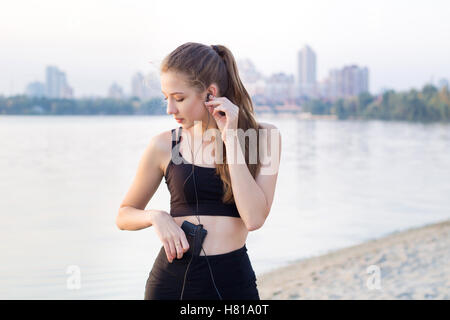 Giovani piuttosto sottile donna fitness sorge sul fiume costa la mattina ascoltando la musica tramite telefono cellulare player earphon Foto Stock