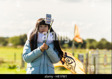 Giovane donna prende un selfie utilizzando un mobile/smart phone e un bastone selfie. Foto Stock