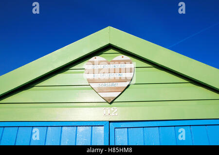 Dipinta di fresco beach hut con un brioso cuore di legno la placca visualizzando il testo "Oh mi piace essere accanto al mare!" Foto Stock