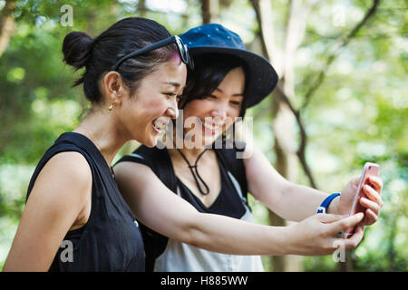 Due giovani donne in piedi in una foresta, prendendo un selfie. Foto Stock