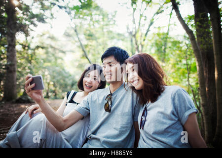 Due giovani donne e un uomo seduto in una foresta, prendendo un selfie. Foto Stock