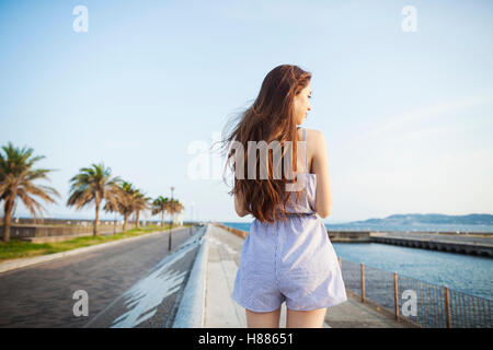 Vista posteriore della giovane donna con lunghi capelli rossi in piedi in uno spazio aperto da una strada lungo la costa. Foto Stock