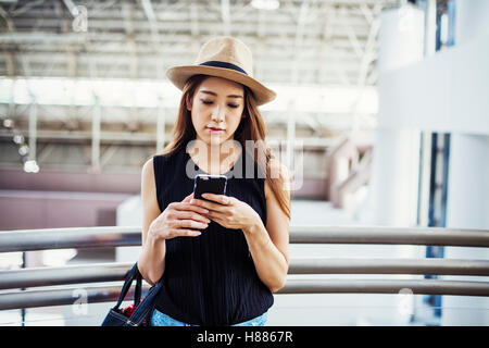 Giovane donna con capelli lunghi marrone, indossando un cappello di Panama, in un centro commerciale, utilizzando un telefono cellulare. Foto Stock