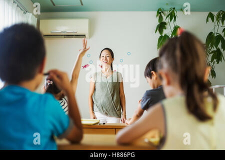 Un gruppo di bambini in una classe, uno con la sua mano pronto a rispondere a una domanda. Foto Stock
