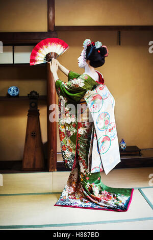 Una Donna vestita con il tradizionale stile geisha, indossando un kimono e OBI, in piedi in una posa classica con ventola sollevato Foto Stock