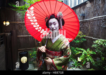 Una Donna vestita in stile geisha, bianco viso trucco con luminosi rossi labbra in possesso di una carta di colore rosso ombrellone. Foto Stock