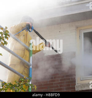 Uomo in giallo tuta antipioggia pulisce la vernice da un muro di mattoni della facciata della casa con la rondella di pressione mentre si sta in piedi sul ponteggio Foto Stock