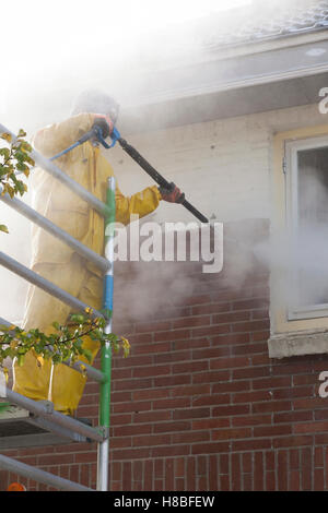 Uomo in giallo tuta antipioggia pulisce la vernice da un muro di mattoni della facciata della casa con la rondella di pressione mentre si sta in piedi sul ponteggio Foto Stock