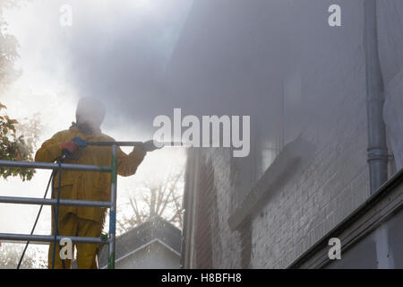 Uomo in giallo tuta antipioggia pulisce la vernice da un muro di mattoni della facciata della casa con la rondella di pressione mentre si sta in piedi sul ponteggio Foto Stock