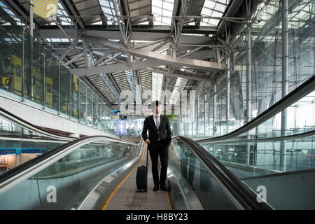 Asian giovane imprenditore con bagagli giù per la scala mobile in aeroporto. Business travel concept. Foto Stock