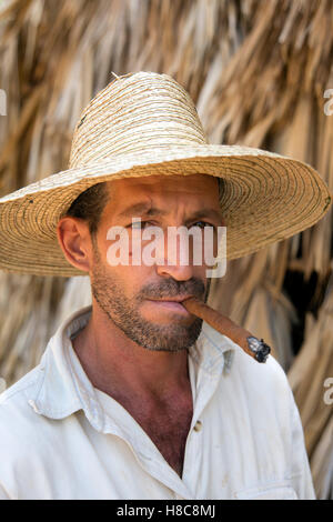 Ritratto verticale di un giovane contadino cubano che indossa un cappello di paglia che fuma un sigaro con la casa di essiccazione del tabacco di paglia sullo sfondo a Vinales Cuba Foto Stock