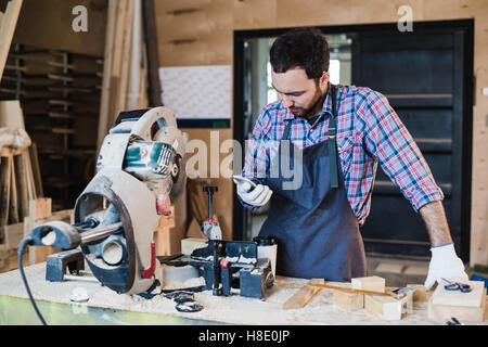 Carpenter texting qualcuno del suo smart phone vicino a sega circolare in un polveroso workshop Foto Stock