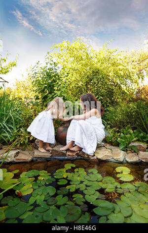Due sorelle in tunica bianca con una brocca in lily pond. Tempo per la famiglia. Foto Stock