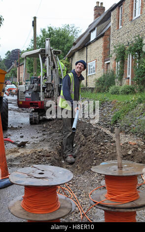 Un grinning workman installazione di cavi per superfast a banda larga in un territorio rurale Oxfordshire village, Regno Unito. Mostra tamburi del cavo ottico. Foto Stock