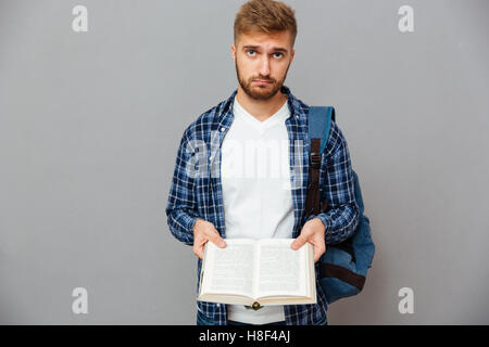 Malinconici uomo barbuto con zaino azienda libro aperto con pagine vuote isolate su uno sfondo grigio Foto Stock