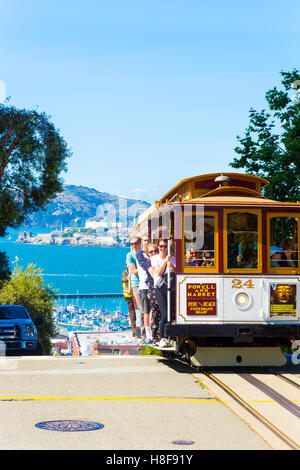 Cavo auto con i turisti che impugnano al di fuori dei corrimano di salita ripida discesa precipizio a picco di Hyde St con vista di Alcatraz SF Foto Stock