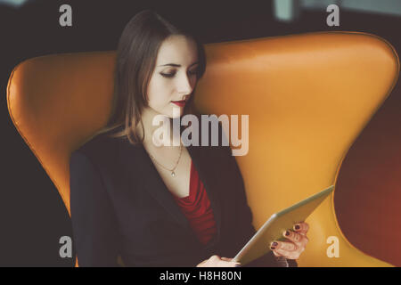 Ritratto di giovane donna di successo imprenditore in un abito rosso e giacca seduta sulla poltrona di colore arancione e lavorando al suo digital Foto Stock