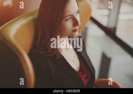 Ritratto di giovane donna di successo imprenditore in un abito rosso e giacca seduta sulla poltrona di colore arancione vicino alla finestra con la riflessione Foto Stock