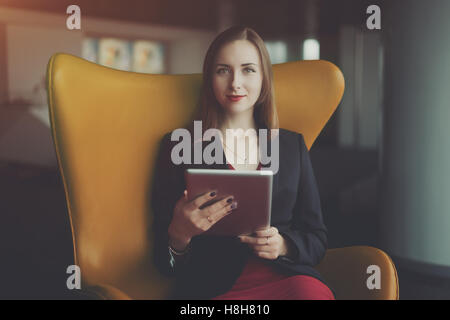 Ritratto di giovane donna di successo imprenditore in un abito rosso e giacca seduta sulla poltrona di colore arancione e lavorando al suo tablet Foto Stock