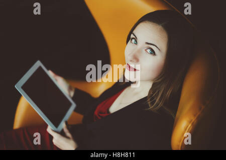Ritratto di giovane donna di successo imprenditore in un abito rosso e giacca seduta sulla poltrona di colore arancione con tavoletta digitale Foto Stock