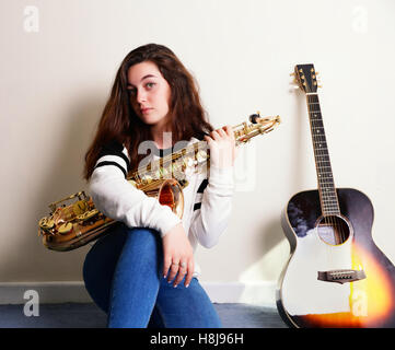 Ragazza adolescente tenendo un sassofono con una chitarra in background Foto Stock