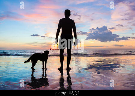 Silhouette di un uomo e di un cane in piedi insieme sulla spiaggia al tramonto e guardando all orizzonte, amicizia tra uomo e animale Foto Stock