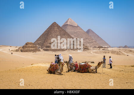 Il Cairo, Egitto i turisti e i driver del cammello con i loro cammelli in appoggio nel deserto con le tre grandi piramidi di Giza in bac Foto Stock