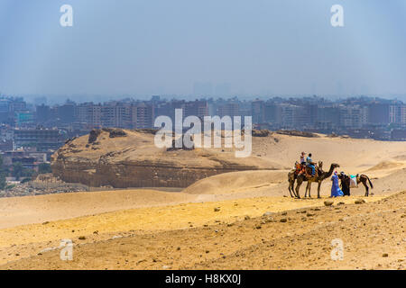 Il Cairo, Egitto Camel driver e i turisti a cavallo di cammelli e cavalli attraverso il deserto con la città del Cairo in background. Foto Stock