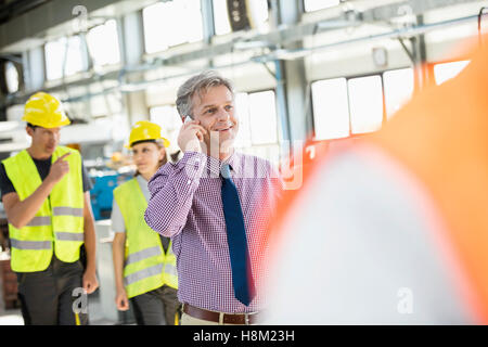 Supervisore maschio parlando al telefono mobile con i lavoratori in background all'industria Foto Stock