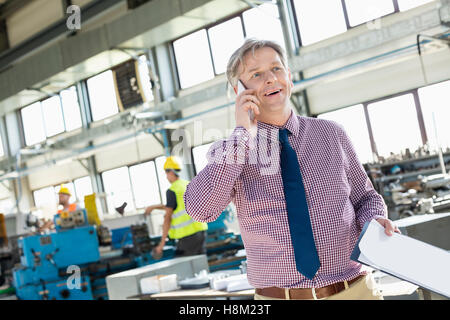 Maschio maturo supervisore con appunti parlando al telefono cellulare nell'industria Foto Stock