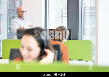 Adulto medio imprenditore in piedi con le braccia incrociate con le colleghe che lavorano in primo piano in ufficio Foto Stock